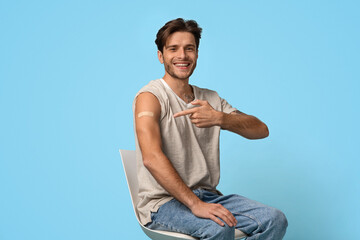 A young man with medium-length hair sits in a chair, smiling as he points to a small bandage on his arm. The bright blue background adds to the cheerful atmosphere.