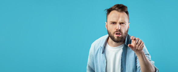 A man with a beard is pointing forward with a serious expression. He wears a casual white t-shirt and a blue jacket, creating a striking contrast against the background.
