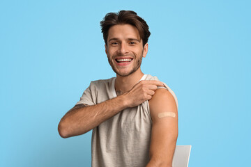 Smiling young man in a casual gray shirt points to a bandage on his upper arm. He appears happy and proud of getting vaccinated, with a light blue background.