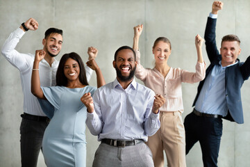 Group of diverse coworkers joyfully celebrates a significant business achievement, displaying excitement and unity with raised fists in a modern office setting.