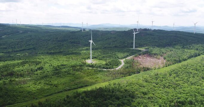 Aerial view of windmills above a forest in Saint-Robert-Bellarmin with a foggy mountain range in the background and partial sunlight. Scenic renewable energy landscape in rural Quebec.