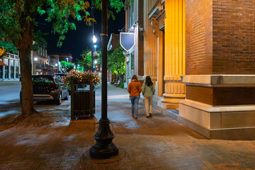 Two young women walk along the brick paved sidewalk at night in the historic downtown district of...