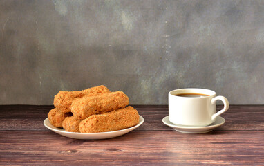 Five fresh eclairs with sprinkles on a plate on a wooden table, next to a cup of coffee.