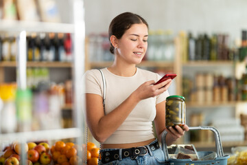 Young woman buyer scanning qr code for pickled cucumbers in jar in grocery store