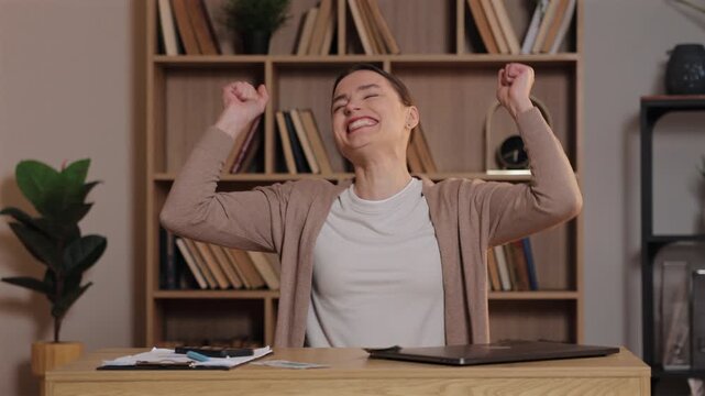 Caucasian woman laughing excitedly throwing cash in air sitting at desk. Female enjoying success scattering money around in joyful mood at home office with laptop and bookshelf in background.