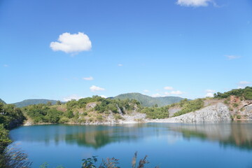Fototapeta premium Blue Lagoon Phuman Khon Kaen: Majestic gray limestone cliff beside a clear turquoise water pond, with perfect reflection under the summer sky