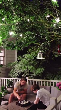 Cozy Summer Night on the Porch &ndash; Father and Son Playing Checkers Beneath Glowing String Lights on Cozy Terrace