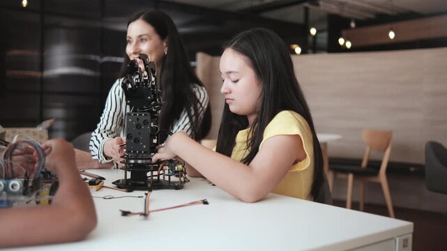 Student girl robotic arms designs and experiments with White female teacher instructing at learning table in primary science classroom, discussing technology knowledge for kids of elementary school.