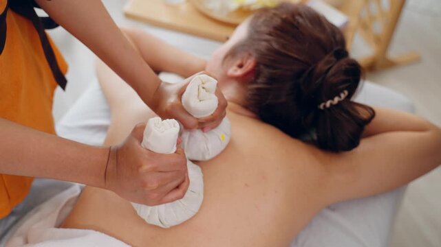 Beautiful woman getting thai hot compress massage with herbal bags. Attractive young girl lying down on massage bed, getting relaxing physiotherapy service from masseuse in the spa beauty clinic.
