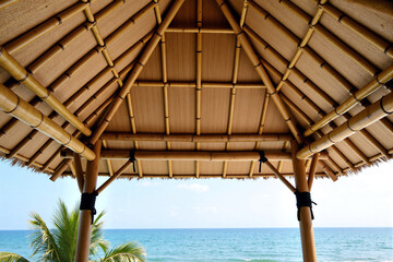 Low angle view of traditional wooden bamboo roof structure of beach gazebo or hut against blue sky and palm trees for tropical summer vacation concept