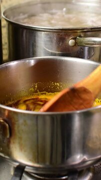 Close-up stainless steel pot with melted pork lard infused with annatto (onoto), being stirred with wooden spoon while a pot simmers in background. Traditional preparation process Venezuelan hallacas.