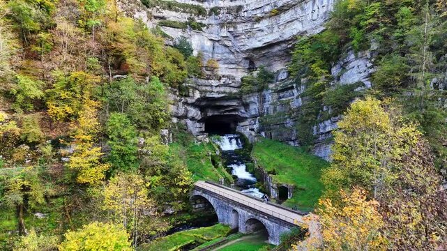 Limestone cliff of Source de la Loue river bursting from cavern and cascades, down stepped waterfall passing underl arched stone road bridge in Ouhans, Doubs, Bourgogne‑Franche‑Comt&eacute;, France aerial