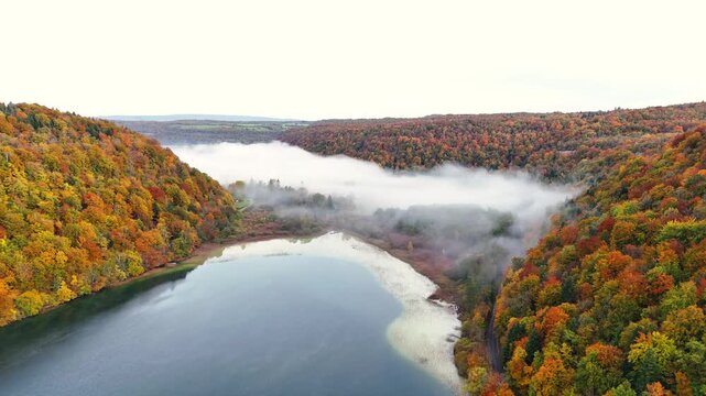 Drone establishing shot captures Lac du Val lake surface, forest surrounds valley walls show autumn foliage colors, thick mist fog hovers low water surface, Jura mountains, Le Herisson, France