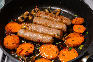 Sausages and sweet potatoes cooking in a skillet on a stove with mushrooms and herbs in a kitchen setting