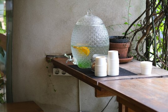 A large glass water dispenser with citrus slices sits on a wooden counter beside stacked paper cups, creating a refreshing self-serve drink station.

