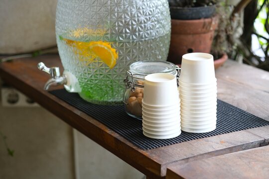 A large glass water dispenser with citrus slices sits on a wooden counter beside stacked paper cups, creating a refreshing self-serve drink station.