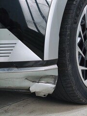 A vertical close-up of a severe accident, impact, or scrape damage to the silver plastic front bumper fender of a car, showing deep black scratches, cracks, and distortion near the wheel