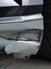 A vertical close-up of a severe accident, impact, or scrape damage to the silver plastic front bumper fender of a car, showing deep black scratches, cracks, and distortion near the wheel