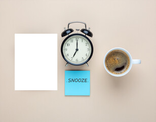 Flat lay of a black alarm clock, a cup of coffee, a blank white paper, and a blue sticky note with "SNOOZE." on a beige background, symbolizing morning routine.