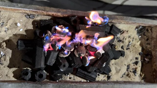 Overhead shot of glowing charcoal briquettes on a bed of sand in a rusty metal grill. Vibrant blue and orange flames dance over the coals, creating intense heat for outdoor BBQ or cooking.