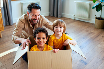Caucasian man smiling while playing with two children, one Caucasian boy and one biracial boy, both sitting inside cardboard box on wooden floor, all looking happy and engaged