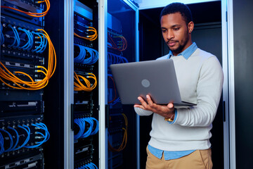 Young adult Black man working in server room holding laptop, managing network cables and equipment, focusing on system maintenance and data center operations, standing near server racks