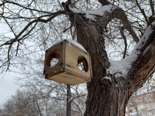A wooden bird feeder covered in snow, hanging from a bare tree branch