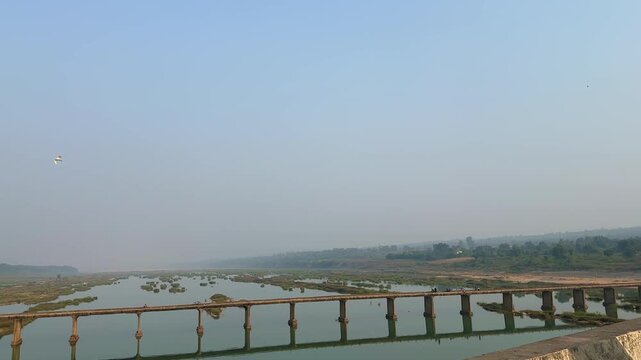 An old concrete bridge spans the calm Son River, its pillars reflected in shallow waters and sand patches, surrounded by open skies and distant green hills.