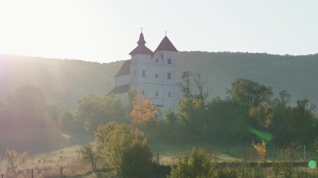 Aerial photo of a white church with red roofs on a hill, in a foggy morning landscape. Herina, Bistrita Nasaud, Romania.