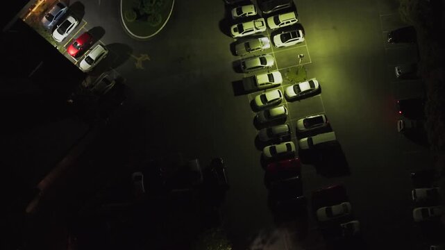 Drone aerial footage of a well-lit parking lot at night, showcasing orderly parked cars and glowing streetlights casting shadows on the pavement. Perfect for urban evening scenes.