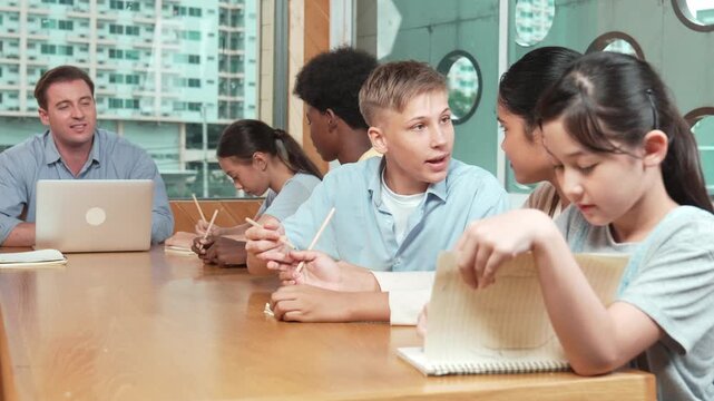 Asian girl laughing at caucasian boy idea while professional teacher teaching about program. Happy diverse student work classwork while sitting at classroom. Creative education concept. Edification