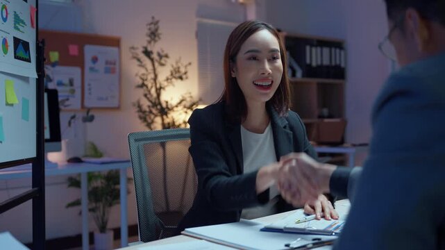 Successful young asian businesswoman shaking hands with a male partner, finalizing a deal in a modern office during the late evening