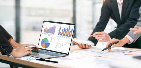 Close-up of three business professionals meeting in an office, focusing on financial charts displayed on a laptop and paper documents.