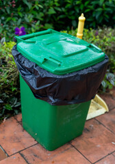 A green outdoor trash bin with a black garbage bag, set against vibrant greenery.