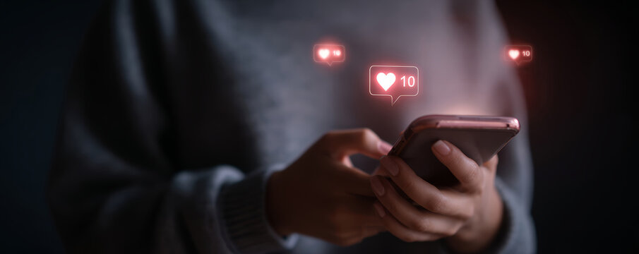 Person holding smartphone with social media like notification icons glowing in dark background - Powered by Adobe