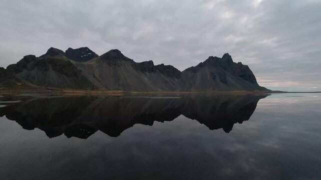 Static wide shot capturing the dramatic, jagged peaks of the Vestrahorn mountain reflected perfectly in the still, mirror-like dark water under a rapidly passing cloudscape.