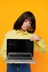 Fototapeta premium A young woman in a yellow shirt holds an open laptop, gazing downward with a pout as bright orange backdrop highlights a moment of tech frustration or focus.