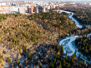 Fototapeta premium Aerial view capturing a winding, snow-covered ski track cutting through a mixed winter forest near urban development in February's golden light