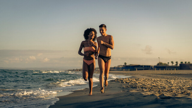 A Joyful Diverse Couple Runs Barefoot Along the Beach at Golden Hour