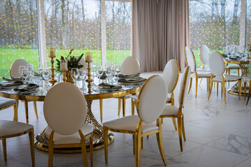 A luxurious banquet hall with round tables set for an elegant event, featuring white chairs, gold accents, and large windows