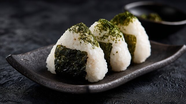 Close up view of Japanese onigiri rice balls seaweed wrapping served on ceramic plate against dark black textured backdrop