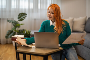 Woman working at home office with laptop, documents, and indoor plants in cozy living room setting