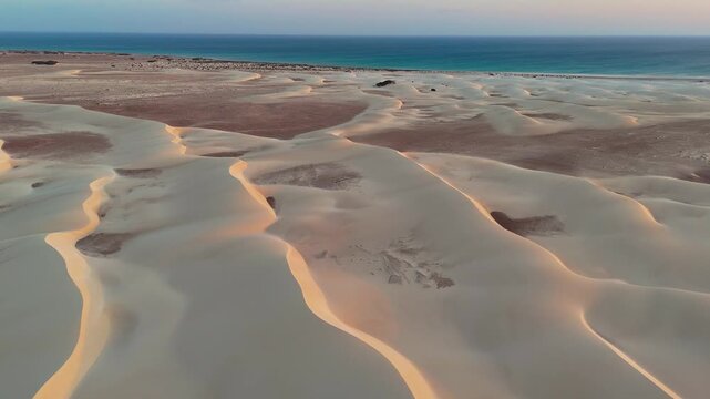 Drone view of flowing desert sand dunes glowing in warm sunset light as they meet a deep blue sea, creating a striking contrast between arid land and ocean horizon. Socotra island, Yemen, AfricaQ