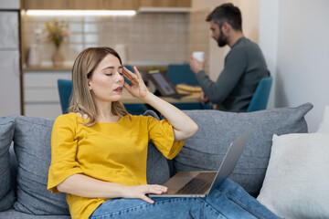 Woman sitting on a couch struggling with a headache while a man works on a laptop in a modern kitchen setting