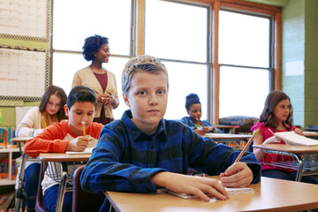 Portrait, boy student and learning in classroom with pencil for knowledge, education and assignment. Child, classmates and teacher in school with notebook for workbook activity, assessment or lesson.