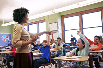 Classroom, teacher and children with hands up, education and students with academic growth or learn. Happy, woman and kids with gesture for answer, knowledge and cognitive development in school