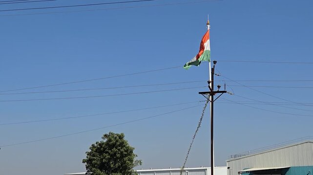 India's vibrant Tiranga flag catches the wind atop a tall mast, overlooking a bustling industrial area with factory buildings, a large truck, and numerous power lines.