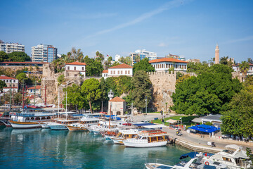Scenic view of Antalya’s old harbor with traditional boats docked along turquoise Mediterranean waters, framed by historic stone city walls, lush greenery, and red-roofed Ottoman-style buildings