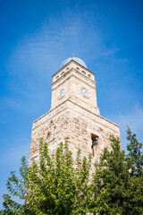 Obraz premium Low-angle view of a historic stone clock tower rising above green trees under a clear blue sky in Turkey. The timeless architecture, weathered limestone texture, and classic clock tower