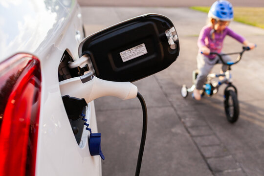 Electric vehicle charging in family home driveway with child riding bike in background